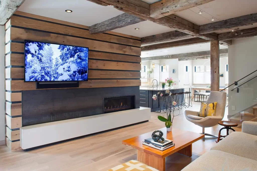 Warm modern living room with wood fireplace surround, exposed beams, and open kitchen connection.