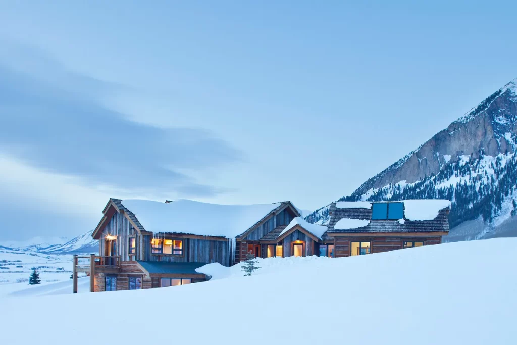 Snow-covered riverfront home in Crested Butte showcasing timber detailing and solar panels under a blue sky.