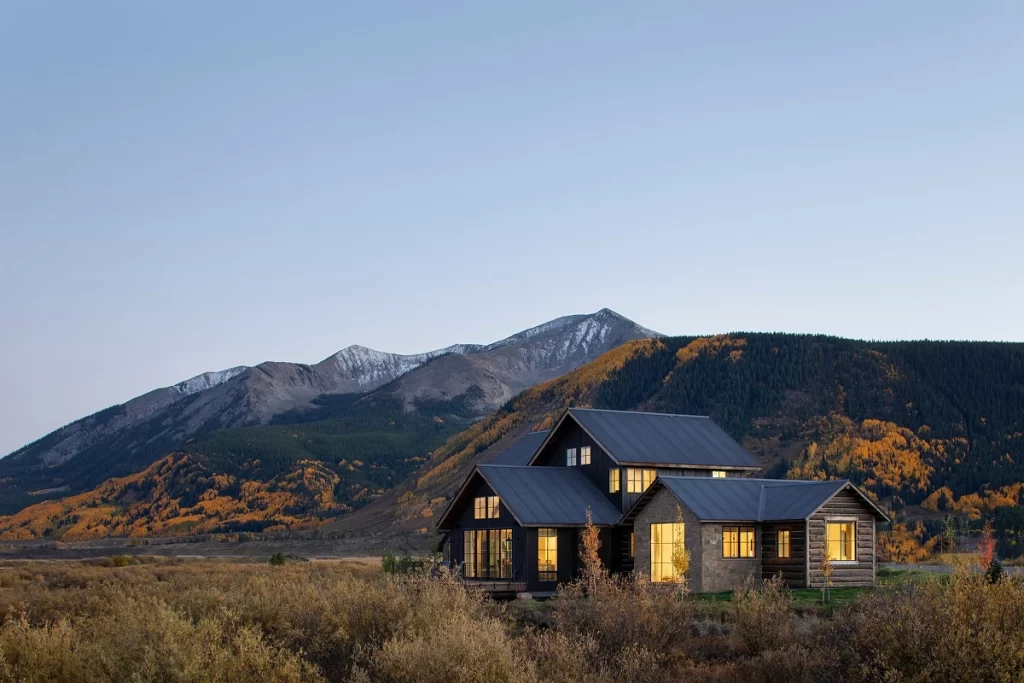 Evening view of a mountain modern home with warm interior light and dramatic peaks in the background.